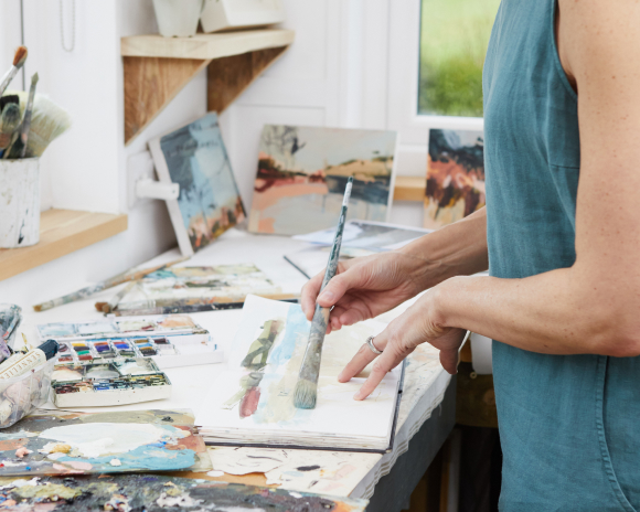 A desk covered in painting supplies and an open notebook and hands holding a paintbrush painting in the notebook