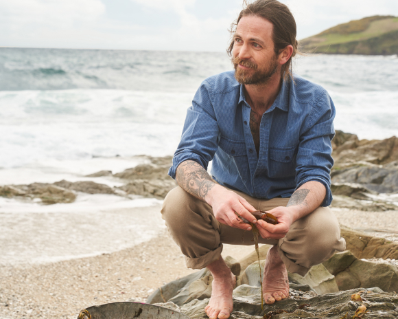 A man wearing a blue shirt and brown shorts is crouched on a rock on a beach