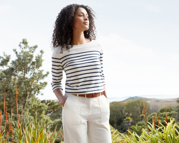 A woman in a field with brown curly hair wearing white trousers and White and navy striped top