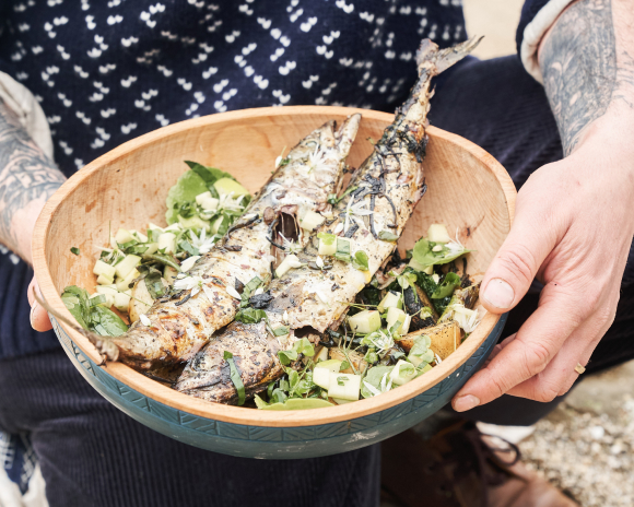 Hands holding a wooden bowl with salad and 2 whole grilled mackerel inside.