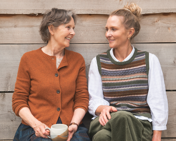 two women smiling at each other, one is wearing an orange cardigan and the other is wearing a white shirt with striped knitted vest