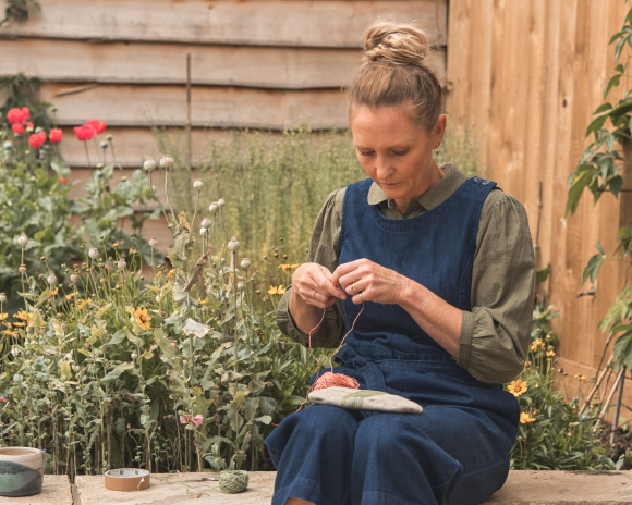 A women wearing a denim jumpsuit with green shirt sat weaving on a bench in front of a bed of flowers. 