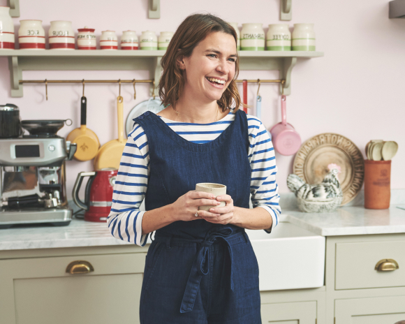 A smiling woman in a striped top and denim dungarees holding a mug in a kitchen.