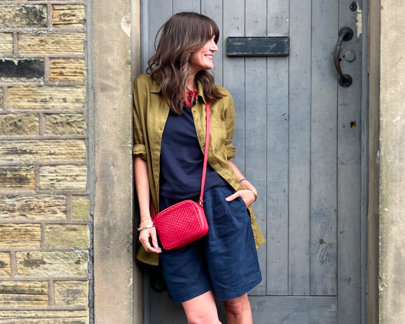 Lady with brown hair looking to the right wearing green shirt and navy shorts with red bag leans in doorway with a pale blue door behind her.