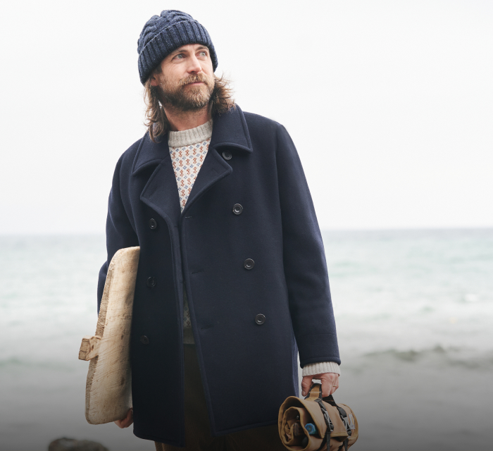 Man wearing Seasalt hat and coat at the beach, holding a chopping board and a tool wrap.