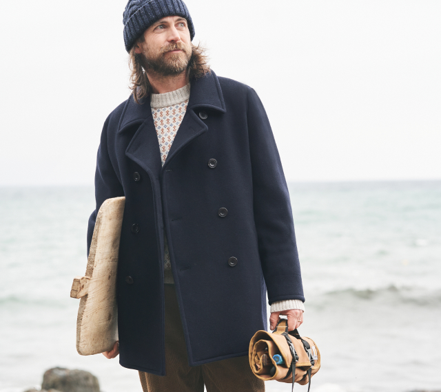Man wearing Seasalt hat and coat at the beach, holding a chopping board and a tool wrap.